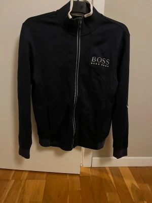 Hugo Boss Ziptröja - Säljer denna ziptröjan från Hugo Boss då den inte kommer till användning längre. Den är i hyfsat bra skick men har tecken på användning. Finns mindre märken på båda ärmarna. Den har även reflex både i loggan på bröstet och på linjerna på ärmarna. 