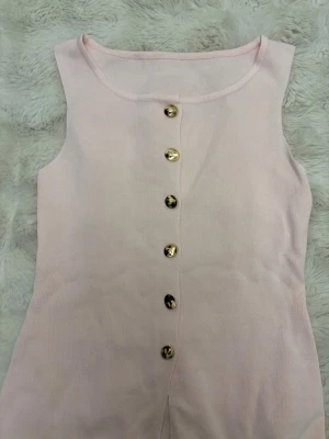 Ljusrosa ärmlös linnetopp med knappar - Supersöt ljusrosa linnetopp med guldfärgade knappar framtill. Toppen är ärmlös och har en rund halsringning. Materialet är mjukt och ribbat, vilket ger en skön känsla mot huden. Perfekt för varma dagar och enkel att styla med jeans eller kjol.