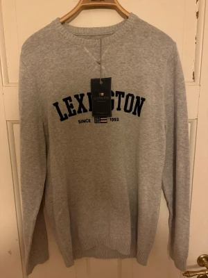 Grå Lexington tröja med tryck - Snygg grå tröja från Lexington med mörkblått tryck på bröstet och ribbade muddar vid ärmslut och nederkant. Rund hals och långärmad modell, perfekt för chill dagar. Materialet är mjukt och känns skönt mot huden.