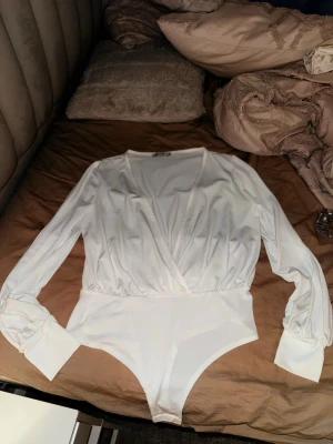 Vit omlott bodysuit från NLY One - Säljer en vit bodysuit från NLY One i storlek S. Modellen har omlott-design med v-ringning, långa ärmar med breda muddar och är tillverkad i ett mjukt, stretchigt syntetmaterial. Perfekt att styla med jeans eller kjol för en clean look.