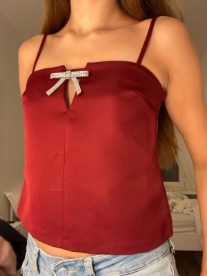 Vinröd satin linnetopp med rosett - Snygg vinröd linnetopp i satin med smala axelband och en glittrig rosett framtill. Toppen har en liten öppning under rosetten som ger en cool detalj. Perfekt att styla med jeans eller kjol för en trendig look.