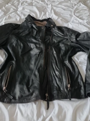 Svart skinnjacka från Levinsky - Svart skinnjacka från Levinsky med coola detaljer som dragkedjor på både fram- och sidofickor. Jackan har hög krage med knapp och är helsvart med snygg passform. Perfekt för dig som vill ha en klassisk men edgy look. I väldigt fin skick storlek 44 men kan passa till 42 också.