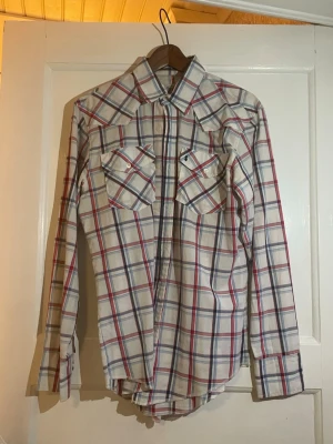 Rutig skjorta från Silver Dollar - Säljer en rutig skjorta från Silver Dollar i storlek L. Skjortan har vit bas med röda och blå rutor, klassisk krage och två bröstfickor med knappar. Tillverkad i bomull, perfekt för dig som gillar en chill och tidlös stil.