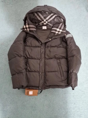 Svart pufferjacka från Burberry - Svart pufferjacka från Burberry med klassiskt rutigt foder i huvan. Jackan har dragkedja och knappar framtill, samt en patch med Burberry London England på ärmen. Perfekt för kalla dagar och har en riktigt snygg streetstil.