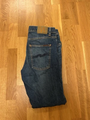 Mörkblå jeans från Nudie - Snygga mörkblå jeans från Nudie med klassisk femficksdesign. Jeansen har kontrastsömmar och Nudie-logga på bakfickan samt läderpatch i midjan. Tillverkade i slitstarkt bomullsmaterial med en autentisk tvättad look. W32 L30