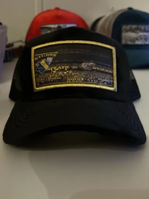 Svart AIK keps med broderad patch - Säljer en svart keps med broderad AIK-patch framtill i gult och blått. Kepsen har mesh på sidorna och justerbar snapback baktill. Insidan har svarta band med texten 'GNAGET'. Perfekt för dig som vill visa ditt AIK-supporterhjärta. Helt ny, inte ens provad.