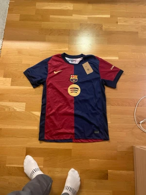 FC Barcelona matchtröja Nike - Säljer en officiell FC Barcelona matchtröja från Nike med klassiska marinblå och vinröda paneler. Tröjan har klubbmärke, Nike-logga och Spotify-sponsor i guld på bröstet. Perfekt för dig som älskar fotboll och vill visa ditt stöd för Barca.