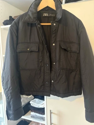 Svart pufferjacka från Zara, M - Svart pufferjacka från Zara med två stora fickor framtill och knappar. Jackan har en klassisk krage och är  i syntetmaterial. Perfekt för dig som gillar en enkel och clean look med lite volym i siluetten.