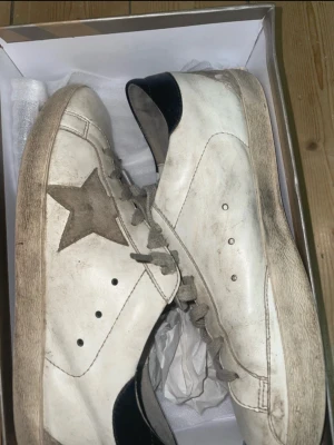 Golden goose skor  - Säljer ett par vita sneakers med grå stjärna på sidan och svarta detaljer vid hälen. Skorna har snörning och platt sula i svart. Tillverkade i skinn med klassisk rund tå och stilren look. Perfekta för dig som gillar streetstyle och vill ha något som sticker ut.