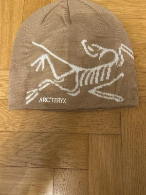 Beige mössa från Arc'teryx - Säljer en beige mössa från Arc'teryx med vit logotyp och skelettmönster på framsidan. Mössan är stickad och har en enkel, rundad form som sitter snyggt på huvudet. Perfekt för kalla dagar och outdoor-aktiviteter.