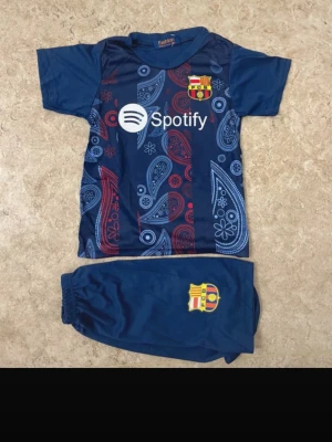 Barça fotbollströja Lamine Yamal #19 - Säljer ett blått FC Barcelona fotbollsställ med Lamine Yamal #19 på ryggen. Tröjan har paisleymönster i rött och blått, samt klubbmärke och Spotify-logga framtill. Matchande blå shorts med FCB-emblem ingår. Perfekt för dig som älskar fotboll och Barça!