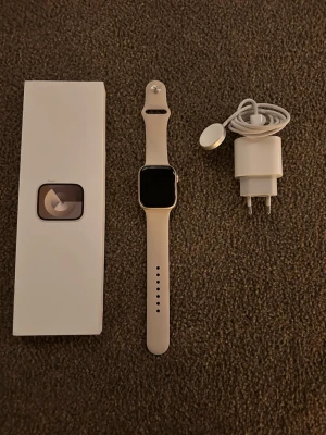 Series 9, starlight aluminum Apple Watch med ljusgrått silikonband - Säljer en Apple Watch med fyrkantig svart display och ljusgrått silikonarmband. Klockan har en stilren design med silverfärgad ram och digital krona på sidan. Levereras med originalförpackning och armbandet är justerbart för perfekt passform.