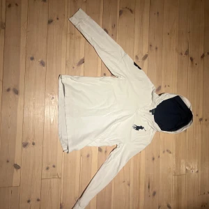 Vit hoodie från Polo Ralph Lauren - Vit hoodie från Polo Ralph Lauren med marinblått foder i huvan, broderad polospelare på bröstet och siffran 3 på ärmen. Tröjan har lång ärm, knappar vid halsen och är tillverkad i bomull med en klassisk, sportig look.