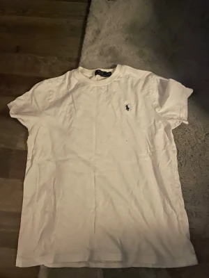 Vit t-shirt från Polo Ralph Lauren - Vit t-shirt från Polo Ralph Lauren med klassisk rund hals och korta ärmar. På bröstet finns den ikoniska broderade loggan i svart. T-shirten är gjord i mjuk bomull och har en normal passform, perfekt för en clean och enkel stil.