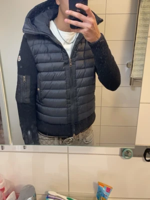 Svart pufferjacka från Moncler - Säljer en svart pufferjacka från Moncler med stickade ärmar och huva. Jackan har dragkedja framtill, fickor med dragkedja på ärmen och Moncler-logga på vänster arm. Perfekt för dig som vill ha en snygg och varm jacka med street vibe.