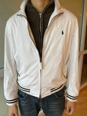 Vintage polo ralph laruen - Snygg vit vindjacka från Polo Ralph Lauren med klassisk broderad logga på bröstet. Jackan har svarta ränder på muddar och midja, dragkedja framtill och sidofickor med zip. Perfekt för dig som gillar vintage kläder 