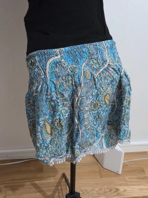 Blå paisleymönstrade shorts Bali - Säljer ett par blå paisleymönstrade shorts från Collection Bali. De har hög midja med resår, lös passform och fina virkade detaljer längst ner. Materialet känns lätt och luftigt, perfekt för varma dagar. Mönstret är färgstarkt med inslag av gult och vitt.