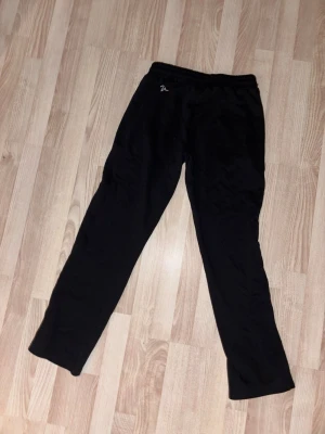 Svarta mjukisbyxor med resår - Svarta mjukisbyxor med elastisk midja och enkel design. Perfekta för chill dagar eller när du vill vara extra bekväm. Liten broderad detalj vid midjan. Mjukt material som känns skönt mot huden. Limiterade byxor går ej o köpa längre