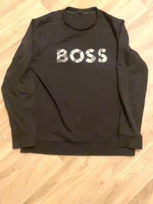 Svart sweatshirt från BOSS, storlek L - Svart sweatshirt från BOSS i storlek L med stort BOSS-tryck i silver på bröstet. Tröjan har rund hals, långa ärmar och ribbade muddar vid ärmslut och nederkant. Perfekt för dig som gillar stilrena och enkla plagg med tydlig logga. Den är L men passar i M också 