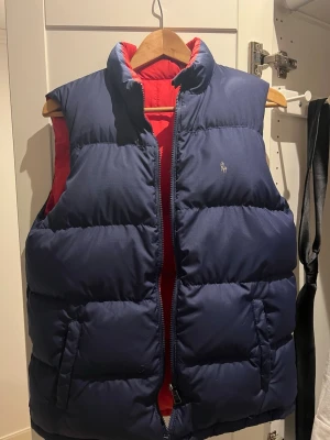 Mörkblå dunväst från Polo Ralph Lauren - Säljer en mörkblå dunväst från Polo Ralph Lauren med röd insida och klassisk logga på bröstet. Västen har hög krage, två fickor med dragkedja och quiltad design. Perfekt att slänga över en hoodie när det är kyligt ute.
