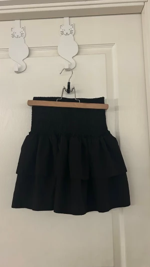 Svart volangkjol med smock - Supersöt svart kortkjol med inbyggd shorts. Bred smock i midjan och dubbla volanger. Kjolen har en tight passform upptill och faller ut i två lager volang, vilket ger en riktigt trendig och lekfull look. Perfekt att styla med en croppad topp eller oversized hoodie.