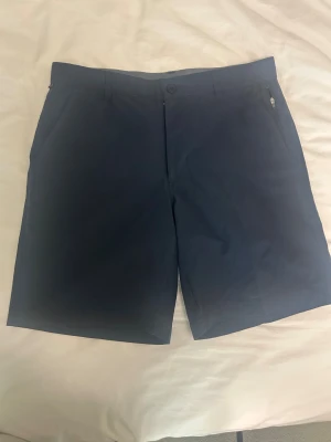 Mörkblå shorts från Swannies - Snygga mörkblå shorts från Swannies med klassisk passform och diskreta fickor på sidorna. Perfekta för sommardagar eller en runda på golfbanan. Tillverkade i ett lätt och bekvämt material som andas. Midjan har en stilren grå insida med texten 'Celebrate Pars'.