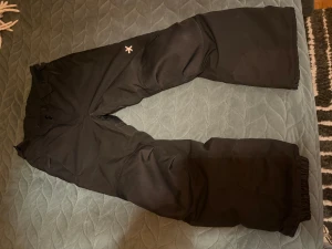 Svarta täcknyxor Everest stl146  - Svarta täckbyxor från Everest med förstärkta partier på knän och bak, samt resår vid benslut. Byxorna har två knappar i midjan och är gjorda i slitstarkt syntetmaterial som tål kyla och snö. Perfekta för vinterns alla äventyr.