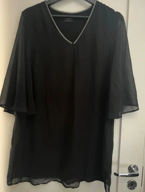 Svart blus med v-ringning - Säljer en svart blus från Red Queen med v-ringad hals och transparenta, vida ärmar. Blusen har en tunn, luftig känsla och en diskret glittrig kant runt halsen som ger en snygg detalj. Perfekt för dig som gillar stilrena och eleganta plagg.