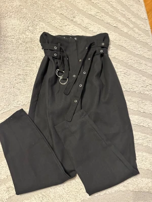 Svarta högmidjade byxor med bälte - Svarta byxor med hög midja och dubbla bälten med metallringar och nitar. Byxorna har rak passform och dragkedja framtill. Materialet är mjukt och känns som bomull eller polyester. Snygga detaljer med öglor och bältesremmar för en edgy look.