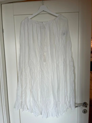 Vit långkjol med tofsdetaljer - Luftig vit långkjol i bohemisk stil med rynkad struktur och knytband med tofsar framtill. Klänningen har en lös passform och är tillverkad i ett tunt, lätt material som passar perfekt för varma dagar.