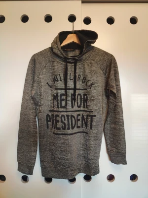 Grå/guld hoodie med tryck och huva - Säljer en gråmelerad hoodie med svart tryck 'I WILL ROCK ME FOR PRESIDENT' på framsidan. Tröjan har huva med svarta snören och långa ärmar. Perfekt för dig som gillar avslappnad stil och vill sticka ut med ett coolt statement.