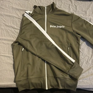 Palm Angels olivgrön track jacket M - Palm Angels track jacket i olivgrönt med vita ränder längs ärmarna och vit dragkedja. Klassisk hög krage och logga på bröstet. Tillverkad i polyester för en sportig vibe. Perfekt för dig som gillar streetwear och vill sticka ut.