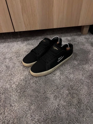 Svarta Saint Laurent sneakers - Svarta sneakers från Saint Laurent med vit sula och guldfärgad logga på plösen. Snygg, stilren design med rund tå och snörning. Materialet är en mix av skinn och textil, vilket ger en lyxig känsla. Perfekt för dig som gillar exklusiva streetwear-vibes.