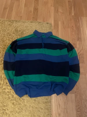 Randig pikétröja i blått, svart och grönt - Säljer en oversized pikétröja med breda blå och gröna ränder. Tröjan har krage och knäppning vid halsen, samt långa ärmar med muddar. Perfekt för dig som gillar retrostil och vill sticka ut lite extra. Materialet känns som mjuk bomull.