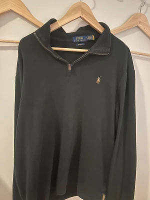 Svart half zip tröja Polo Ralph Lauren - Svart långärmad tröja från Polo Ralph Lauren med klassisk krage och half zip-dragkedja. Tröjan har ett litet broderat Polo-logomärke på bröstet och är tillverkad i mjuk bomull. Perfekt för en chill och stilren look. Tveka inte på att höra av dig om du har eventuella fundering kris pris, kvalité eller annat!