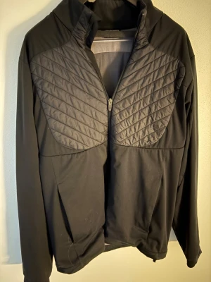 Svart quiltad vindjacka Galvin Green - Svart vindjacka från Galvin Green med quiltade partier fram och bak. Jackan har dragkedja, hög krage och lång ärm. Materialet är syntet och polyester, perfekt för dig som gillar sportig och clean stil. Diskret logga i nacken och snygg passform. Storlek: L passar M