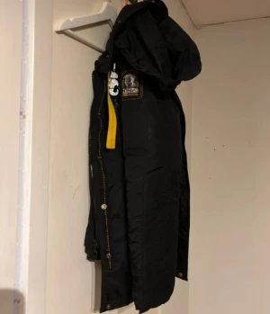 Svart Parajumpers parkajacka med huva - Svart parkajacka från Parajumpers med huva, coolt gult spänne vid kragen och flera fickor med dragkedja. Jackan har patch på ärmen och snygga detaljer i orange och gult. Perfekt för dig som vill ha en stilren och funktionell jacka med streetkänsla.