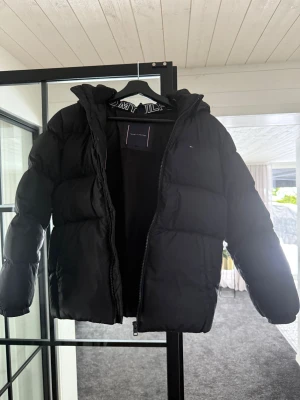 Svart pufferjacka från Tommy Hilfiger - Svart pufferjacka från Tommy Hilfiger med huva och diskret logga på bröstet. Jackan har dragkedja framtill, elastiska muddar och en klassisk pufferlook som håller dig varm. Perfekt för kalla dagar och snygg till streetwear-stilen.