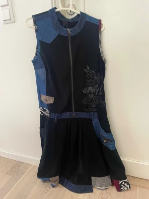 Desigual långklänning med patchwork - Unik långklänning från Desigual med cool patchwork-design i svart, blått och detaljer i grått, rött och mönstrat tyg. Klänningen är ärmlös, har dragkedja framtill och broderade detaljer. Flera fickor och mixade material ger en kreativ vibe.