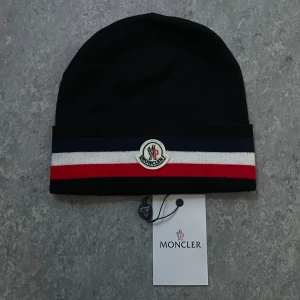 Svart Moncler mössa med ränder - Svart mössa från Moncler med breda randiga detaljer i marinblått, vitt och rött vid kanten. Framtill sitter Moncler-loggan broderad. Snygg och stilren design som lyfter din look, perfekt för kalla dagar. Materialet är mjukt och bekvämt.