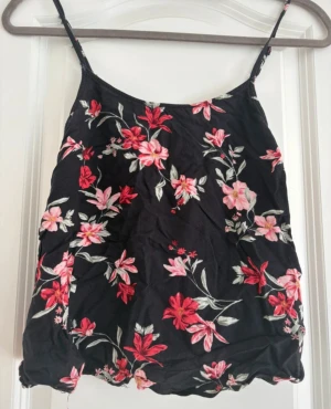 Svart blommig linnetopp från Divided - Svart linnetopp från Divided med tunna axelband och ett färgstarkt blommigt mönster i rött, rosa och vitt. Toppen har en lätt och luftig känsla, perfekt för varma dagar. Materialet är mjukt och bekvämt bomull. Snygg att matcha med jeans eller kjol.