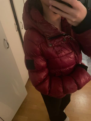 Röd pufferjacka från Superdry - Säljer en röd, glansig pufferjacka från Superdry med huva och svart logopatch på ärmen. Jackan har dragkedja, knappar och är quiltad för extra volym. Perfekt för kalla dagar och har en riktigt snygg streetstil.