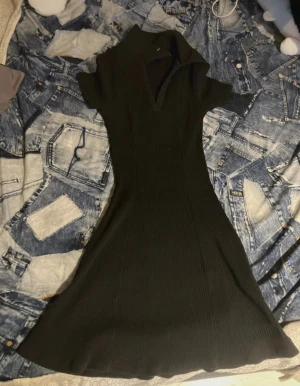 Svart ribbad klänning från H&M - Svart ribbad klänning från H&M med korta ärmar och v-ringning. Klänningen har en lätt utställd form och krage, vilket ger en snygg och modern siluett. Perfekt för dig som gillar stilrena och enkla plagg med en twist.