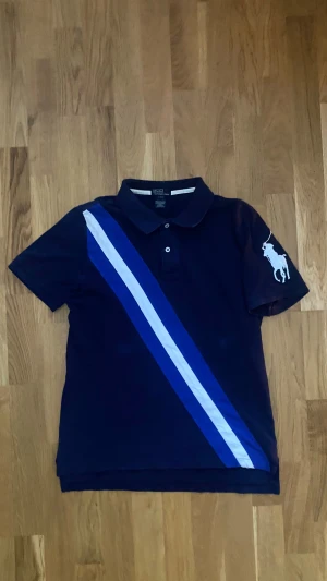 Mörkblå Polo Ralph Lauren piké  - Mörkblå pikétröja från Polo Ralph Lauren i bomull, med klassisk krage och knappar. Snygga diagonala ränder i blått och vitt framtill samt stor vit broderad polospelare på ärmen. Perfekt för dig som gillar sportig och stilren look. Storlek Barn XL men passar herr S, kan bevisa vid önskemål! Vid fler frågor/funderingar tveka inte på att skriva!