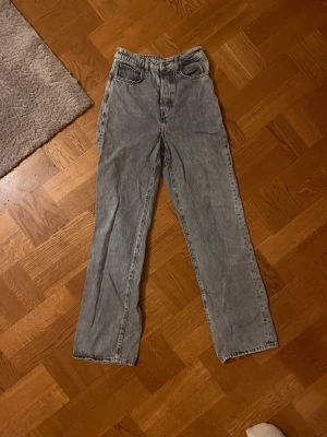 Gråa wide jeans med hög midja - Säljer ett par gråa jeans med snygg tvätt och raka, breda ben. Modellen har hög midja och klassisk knappgylf med flera silverfärgade knappar. Jeansen har fem fickor och bälteshällor, perfekta för en chill och trendig look.