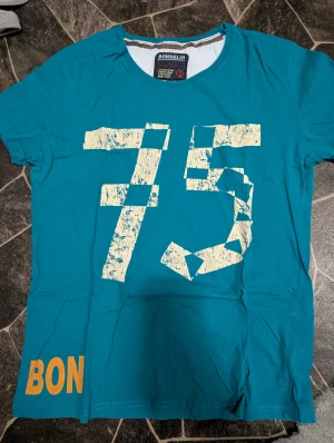 Turkos t-shirt med tryck från Bondelid Stl M - Snygg turkos t-shirt från Bondelid i bomull med stort, slitet tryck av siffran 75 i vitt på framsidan och orange text nere vid kanten. Klassisk rund hals och korta ärmar. Perfekt för dig som gillar sportig och avslappnad stil.