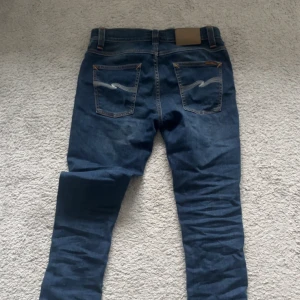 Blå jeans från Nudie Jeans, storlek 30/32 - Snygga blå jeans från Nudie Jeans med klassisk femficksdesign och kontrastsömmar. Modellen har raka ben och subtila slitningar framtill. Bakfickorna har Nudie Jeans signaturbroderi. Passar dig som gillar stilrena och tidlösa jeans.