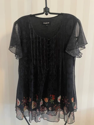 Svart mönstrad topp Desigual M - Svart topp från Desigual med transparenta, korta volangärmar och knappar framtill. Toppen har ett diskret mönster över hela tyget och blommiga detaljer längs nederkanten. Materialet är lätt och meshigt, perfekt för dig som gillar unika prints och detaljer.