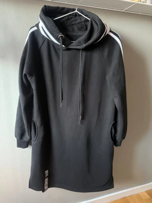 Svart hoodieklänning Real Homegirls - Svart hoodieklänning från Real Homegirls med vita ränder längs ärmarna och stor huva. Klänningen har långa ärmar, fickor i sidorna och snörning i halsen. Mjuk och skön bomullskänsla, perfekt för chill dagar. Logga nertill på framsidan.