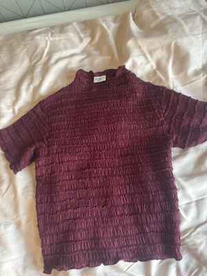 Vinröd smockad topp från Stories XS - Säljer en vinröd smockad topp från Stories i storlek XS. Toppen har korta ärmar, hög hals och är gjord i ett stretchigt material med rynkad struktur. Perfekt för dig som gillar en unik och trendig look. Aldrig använd 